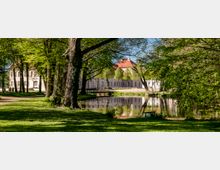 Tree-lined park landscape with a calm pond reflecting surrounding greenery, and a pale historic manor or palace with red-tiled roofs in the background. A grassy path and large mature trees frame the water’s edge.