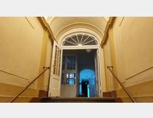 Treppenaufgang in einem historischen Gebäude mit weiß gestrichenen Wänden, verzierten Bögen und offenen Doppeltüren. Dahinter sind weitere Glastüren und ein Flur mit blauer Beleuchtung sichtbar.