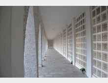 Langer überdachter Korridor mit weißen, grob verputzten Bögen auf der linken Seite und Wänden mit rechteckigen Gedenktafeln auf der rechten Seite, die Namen und Jahreszahlen tragen. Der Boden besteht aus grauen Steinplatten, und am unteren Rand der Tafeln stehen vereinzelt Blumen.