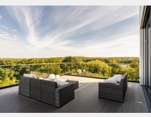 Große Terrasse mit Holzboden, modernen grauen Rattan-Sitzmöbeln und Glasgeländer, umgeben von einer weiten Aussicht auf einen Wald und einen klaren Himmel. Im Hintergrund ist teilweise ein Fluss zu erkennen.