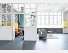 Heller Innenraum mit großen Fenstern, die Tageslicht hereinlassen. Links ein Bereich mit floral gemusterten Wänden, einer orangefarbenen Ledercouch und bunten Dekorationen; rechts ein Bereich mit grauen Sitzmöbeln auf einem Teppich vor einer Heizkörperreihe.