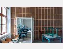 Ein leerer Raum mit großen Fenstern auf der linken Seite und einer mit Holz vertäfelten Wand. Durch eine geöffnete Tür ist ein anderer Raum sichtbar, in dem gestapelte blaue Stühle und Tische zu sehen sind.