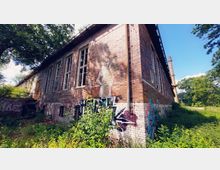 Backsteingebäude mit hohen Fenstern und Graffiti an der Fassade, umgeben von überwuchertem Grün. Im Hintergrund ist ein Schornstein sowie Wald und Himmel zu sehen.
