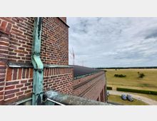 Backsteinfassade eines historischen Gebäudes mit grüner Kupferregenrinne, daneben ein Ziegeldach. Im Hintergrund eine flache, bewachsene Landschaft mit Antennenstrukturen und geparkten Autos auf einem kleinen Hof.