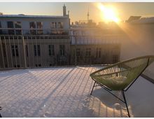 Eine verschneite Dachterrasse mit Metallgeländer, im Vordergrund ein grüner Stuhl mit Eisschicht. Im Hintergrund sind die Gebäude einer Stadt bei Sonnenuntergang zu sehen, mit dem Fernsehturm im Licht der tiefstehenden Sonne.