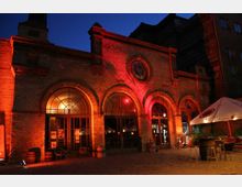 Backstein-Gebäude mit großen Rundbogenfenstern und einem kreisförmigen Fenster im oberen Bereich, beleuchtet von roten Lichtern bei Nacht. Im Vordergrund stehen Tische, Stühle und ein Sonnenschirm auf einem gepflasterten Platz.