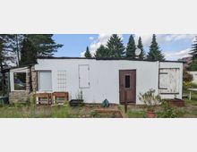 Ein niedriger, länglicher Nebengebäudebau mit hellgrauer Holzverkleidung, Flachdach und mehreren Türen und Fenstern, teils mit Brettern verschlossen. Davor stehen Pflanzkästen und Gartengeräte; im Hintergrund sind hohe Nadelbäume und ein wolkiger Himmel zu sehen.