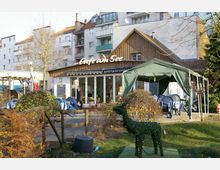 Außenbereich eines Cafés mit dem Schriftzug „Café am See“ auf einem Gebäude mit Holzfassade, großen Glasfenstern und einem grünen Pavillon mit Tischen und Stühlen. Im Vordergrund befinden sich Pflanzen und eine dekorative Figur in Form eines Rentiers aus Grünpflanzen.