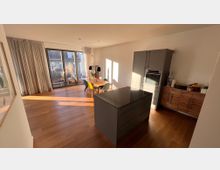 Offener Wohnbereich mit Holzboden, moderner Einbauküche in Grau mit einer zentralen Kücheninsel, einem Esstisch mit bunten Stühlen sowie großen Fensterfronten, die auf einen Balkon mit Pflanzen hinaus führen. Die Wand ist in Weiß gehalten, und das Tageslicht fällt durch die Fenster in den Raum.