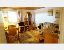 Wohnzimmer mit Holzmöbeln und gemütlicher Einrichtung. Es gibt ein Sofa mit Kissen, einen Couchtisch aus Korbgeflecht, einen Fernseher auf einem Holzschrank sowie eine Vitrine; zwei Fenster sorgen für Tageslicht, daneben hängt eine Tischleuchte an der Decke.