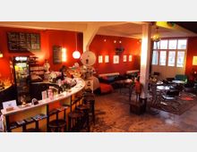 Gemütliches Café mit roten Wänden, einer geschwungenen Theke und schwarzen Barhockern. Im Raum befinden sich kleine Sitzgruppen mit Stühlen und Tischen, eine große Fensterfront sorgt für Tageslicht, und leuchtende Dekoelemente hängen von der Decke.