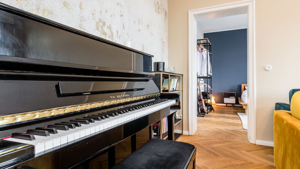 Innenraum einer Wohnung mit schwarzem Klavier im Vordergrund vor einer hellen, teils abgenutzten Wand und Fischgrätparkettboden. Durch einen weißen Türrahmen blickt man in ein angrenzendes Zimmer mit dunkelblauer Wand, Kleiderständer und niedrigem Bett.