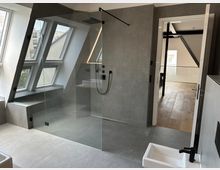 Modernes Badezimmer mit bodengleicher, verglaster Walk-in-Dusche vor einer grauen Fliesenwand und schwarzen Armaturen. Links sorgen große Dachfenster mit Sitzbank für Tageslicht, rechts führt eine offene Tür in einen Flur mit Holzfußboden.
