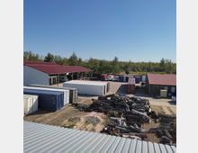 Mehrere Container und Holzstapel auf einem Außengelände mit zwei überdachten Lagergebäuden mit roten Dächern; im Hintergrund sind Bäume unter einem klaren blauen Himmel zu sehen.