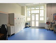 Ein Krankenhausraum mit blauen Böden, weißen Schränken entlang der linken Wand und medizinischen Geräten auf der rechten Seite. Große Fenster und eine Tür im Hintergrund lassen Tageslicht herein.