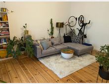 Wohnzimmer mit Holzboden, grauem Ecksofa, einem kleinen runden Couchtisch und mehreren Zimmerpflanzen. An der weißen Wand stehen ein Bücherregal und zwei an der Wand befestigte Fahrräder; eine Stehlampe befindet sich daneben.