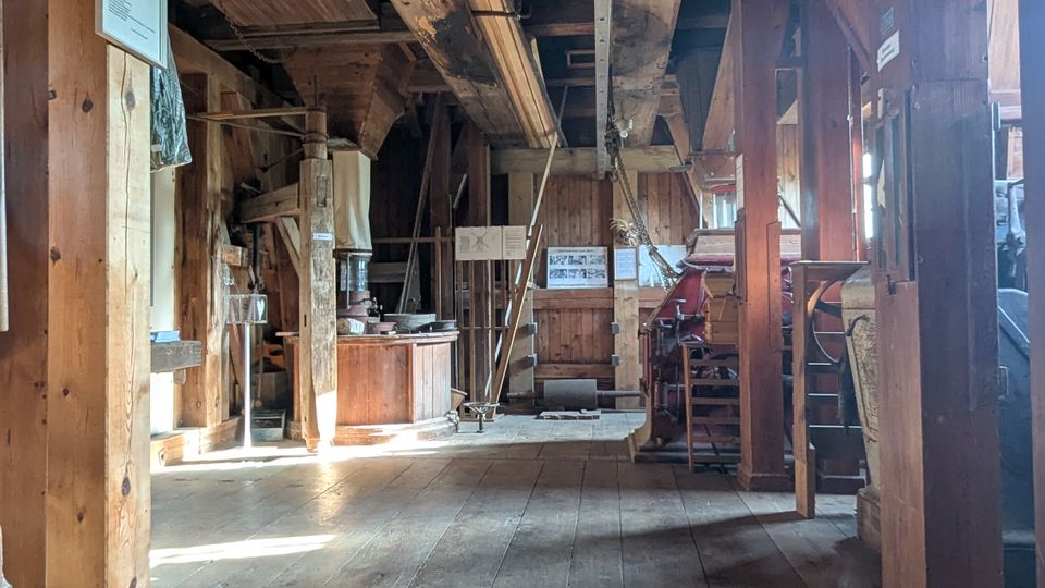Innenraum eines historischen Holzgebäudes mit freiliegenden Deckenbalken, breiten Holzdielen und mehreren Stützpfosten. Entlang der Wände stehen alte Maschinen und Ausstellungsstücke, ergänzt durch Informationstafeln; Tageslicht fällt durch seitliche Fenster ein.