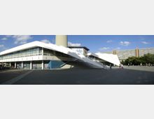 Modernes Gebäude mit einer weißen, schrägen Dachkonstruktion und breiten Treppenstufen, umgeben von einem gepflasterten Platz. Im Hintergrund sind ein hoher Fernsehturm und ein langes Wohngebäude mit Balkonen zu sehen.