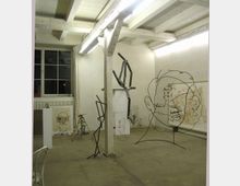 Industrieähnlicher Atelierraum mit rauem Betonboden und Holzbalkendecke, beleuchtet durch Neonröhren. Der Raum enthält moderne Skulpturen aus Metall und Draht sowie Zeichnungen und Gemälde, teils auf dem Boden stehend und teils an die Wände gelehnt.