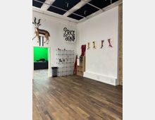 Interior of a studio or shop space with white walls, dark wood floors, and an exposed ceiling grid, featuring a “Price & Son” wall sign, a mounted antelope figure, and shoe-themed artwork. A doorway opens to a room with a green screen, and the wall area includes modular storage cubes and small shelving.