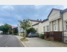 Straßenszene mit historisch anmutenden Wohnhäusern, Gehweg und einer kurvigen Fahrbahn mit Straßenbahnschienen. Ein Auto parkt vor einem Haus mit Säulen, und vereinzelt stehen Bäume entlang des Gehwegs.
