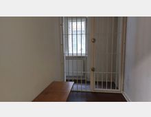 Ein schmaler Raum mit weiß verputzten Wänden, einem hölzernen Sitzbereich links und einer Metallgittertür vor einem vergitterten Fenster. Hinter dem Fenster ist ein Heizkörper sichtbar.