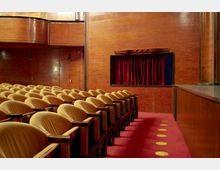 Innenraum eines Theatersaals mit Sitzreihen aus gepolsterten, beige-gelben Stühlen und Armlehnen aus Holz, umgeben von holzvertäfelten Wänden. Ein kleiner Bühnenbereich mit roten Vorhängen ist an der Wand sichtbar, der Boden ist mit einem roten Teppich ausgelegt.