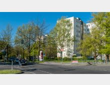 Wohngebiet mit mehrstöckigen Wohngebäuden im Hintergrund, umgeben von Bäumen und Grünflächen. Im Vordergrund eine Kreuzung mit Fußweg, Fahrrädern, Autos, Straßenlaternen und einer Litfaßsäule.