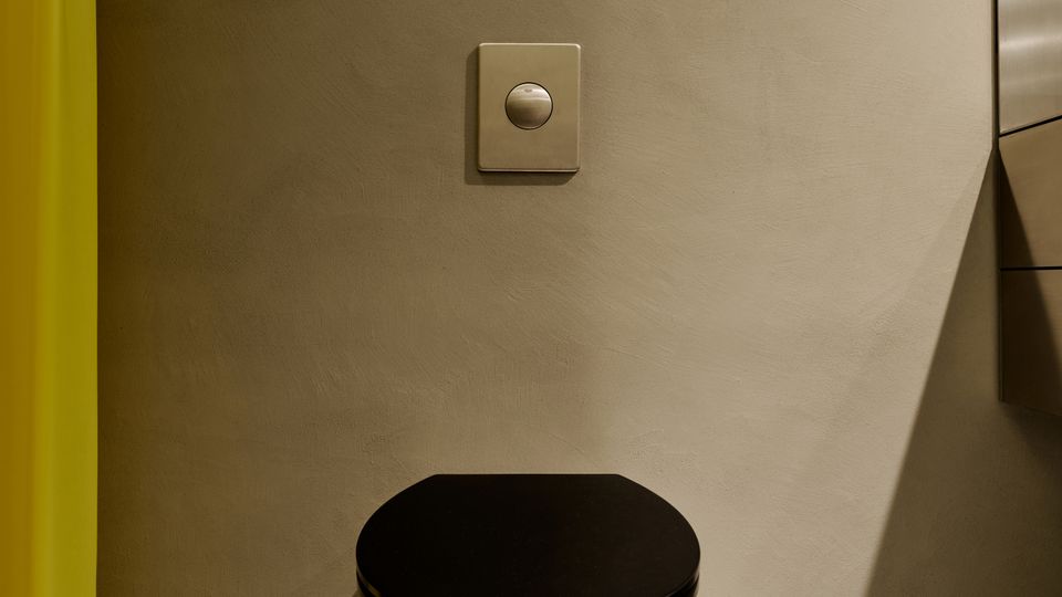 Minimalistischer Innenraum mit glatter grauer Wand, mittig montiertem Druckknopf und einem kleinen zylindrischen Metallhocker mit schwarzer Sitzfläche auf einem hell gesprenkelten Boden. Links ist ein gelber Vorhang zu sehen, rechts eine spiegelnde Fläche mit Leuchte.