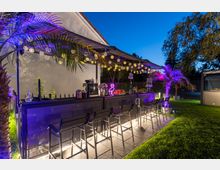 Beleuchtete Outdoor-Bar im Garten mit Hockern, umgeben von grüner Rasenfläche und dekorativen Palmen. Die Bar ist mit Lichterketten und bunten LEDs geschmückt, und auf der Theke stehen Getränke und Dekorationselemente.