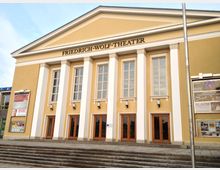 Außenansicht des Friedrich-Wolf-Theaters, einem gelb-beigen Gebäude mit klassizistischen Elementen, darunter vier weiße Säulen und große Fenster. Eingangsbereich mit Holztüren, flankiert von Treppen und Plakaten für Veranstaltungen.