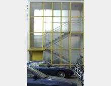 Modernes Gebäude mit einer großen Glasfassade, unterteilt durch gelbe Metallrahmen, die eine Treppe im Inneren sichtbar machen. Im Vordergrund stehen parkende blaue Autos und Fahrräder.