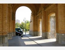 Gewölbter Durchgang aus gelben Backsteinen mit verzierten Säulen und Bögen, flankiert von Pflastersteinen und schattigen Gehwegen. Im Hintergrund sind geparkte Fahrzeuge, Bäume und moderne Gebäude zu sehen.