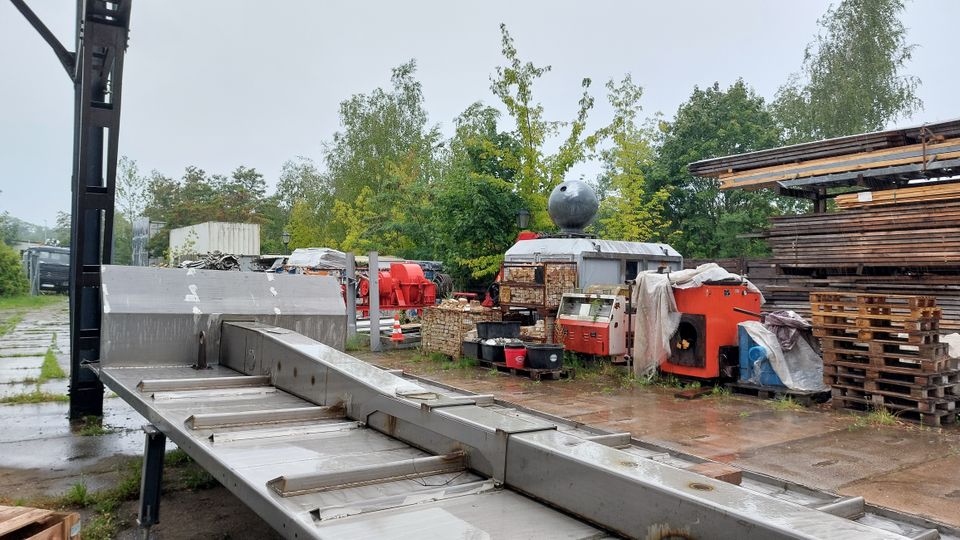 Ein regnerischer Außenbereich mit verschiedenen Maschinen, Holzstapeln und Metallstrukturen. Im Hintergrund stehen Bäume und Container, während der Boden aus Betonplatten besteht.