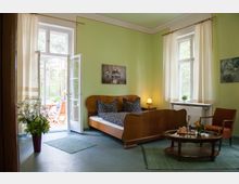 Ein gemütliches Schlafzimmer mit hellgrün gestrichenen Wänden, einem Holzbett mit gemusterten Kissen, einem kleinen Beistelltisch und einem Sessel. Ein Fenster und eine geöffnete Glastür führen auf eine Terrasse mit Blick auf einen bewaldeten Außenbereich.