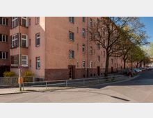 Mehrstöckiges Wohngebäude mit hellrosa Putzfassade und einer unteren Reihe aus rotem Klinkerstein, gelegen an einer Straßenecke. Im Vordergrund stehen Bäume, ein Straßenschild sowie parkende Autos entlang des Gehwegs.