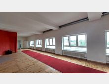 Ein leerer, heller Raum mit Holzboden und weißen Wänden, durchbrochen von einer roten Wand links. Große Fenster auf der rechten Seite lassen Tageslicht herein, darunter Heizkörper; ein roter Teppich verläuft mittig durch den Raum.