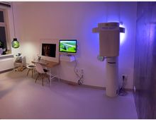 Ein minimalistisch eingerichteter Raum mit einem kleinen Holztisch, zwei weißen Stühlen und einem Computer an der Wand, daneben ein beleuchtetes medizinisches Gerät. Links ein Fenster mit einer kleinen Pflanze und rechts eine Wand mit Metallverkleidung.