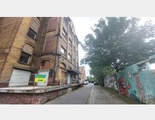 Backstein-Gebäude mit mehreren Etagen und weißen Fensterrahmen, daneben ein mit Graffiti bemalter Holzzaun und ein gepflasterter Weg mit geparkten Autos. Im Hintergrund befindet sich eine Baumreihe.