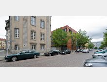 Straßenszene mit einem historischen Gebäude aus hellem Putz und Fensterrahmen aus Stein auf der linken Seite sowie einem modernen Gebäude mit rotbrauner Fassade und großen Fenstern im Hintergrund. Entlang der gepflasterten Straße stehen geparkte Autos, daneben säumen Bäume den Gehweg.