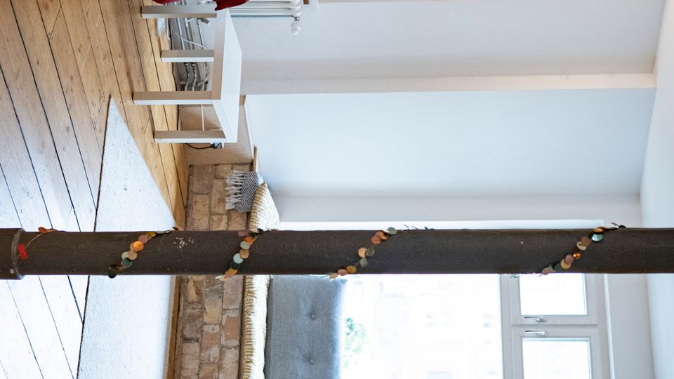 Heller Wohnraum mit offener Küchenzeile links, Holzfußboden und weiß verputzten sowie teilweise freiliegenden Ziegelwänden. Vor großen Fenstern befindet sich eine gepolsterte Sitzbank auf gemauerter Basis, in der Mitte steht eine dunkle Stütze; ein Teppich und ein kleiner Beistelltisch ergänzen den Raum.