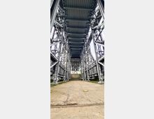 Blick auf das Innere eines massiven Hebewerks mit sichtbarer Stahlkonstruktion, symmetrischen Stahlträgern und Betonboden. Durch die geöffnete Struktur ist im Hintergrund ein heller Außenbereich erkennbar.