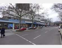 Straßenszene mit einem Einkaufszentrum auf der rechten Seite, das mehrere Geschäfte wie einen Friseursalon, dm und Edeka beherbergt. Im Vordergrund sind Gehwege, parkende Autos und Bäume ohne Laub, während die Straße mit mehreren Fahrspuren sichtbar ist.