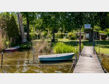 Holzsteg mit Geländer führt zu einem gepflegten Garten mit Rasen, Bäumen und kleinen Hütten; im Vordergrund befinden sich zwei Ruderboote, eines davon im Wasser festgemacht.