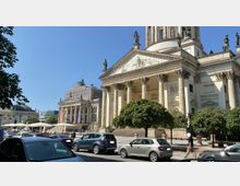 Straßenszene mit Blick auf den Französischen Dom am Gendarmenmarkt in Berlin, einem historischen Gebäude mit Säulenfassade und Statuen. Im Hintergrund sind das Konzerthaus Berlin und weitere historische Bauwerke zu sehen, während im Vordergrund parkende Autos und Fußgänger den Alltag widerspiegeln.