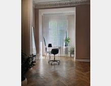 Heller Raum mit Stuckdecke, großen Fenstern und transparenten Vorhängen, ausgestattet mit einem schwarzen Bürostuhl, einem kleinen runden Tisch und Zimmerpflanzen. Der Holzboden ist im Fischgrätmuster verlegt, und die Einrichtung wirkt schlicht und modern.