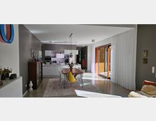 Offener Wohn- und Essbereich in einem modernen Haus mit Küche im Hintergrund. Der Raum verfügt über große Schiebetüren mit Holzrahmen, die viel Tageslicht hereinlassen, einen Esstisch mit mehreren Stühlen, helle Wände, einen Teppich und schlichte Dekorationselemente.