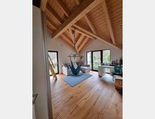 Dachstudio mit offenem, schrägem Holzgebälk, holzfarbenem Boden und großen Fenstern, die auf einen Balkon führen. Im Raum befinden sich eine Hängematte, zwei Sessel, ein Teppich, ein Schreibtisch und eine Holzleiter.