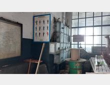 Ein industrieller Raum mit einer großen Fensterwand, mehreren grauen Sicherungskästen an der Wand und einem alten Plakat mit Sicherheitsanweisungen für Eisenbahner. Im Vordergrund befinden sich Werkzeuge, ein grün lackiertes Fass und ein Kabeltrommelhalter.