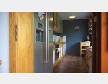 Eine schmale Küche mit braunen Fliesen, Holzarbeitsflächen und weißen Schrankfronten. Ein großer silberner Kühlschrank steht links neben einer Arbeitsfläche, während rechts Regale und ein kleiner Holzschrank platziert sind; eine Wand ist dunkelgrau gestrichen.